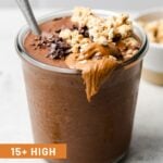 Chocolate protein smoothie in a glass jar topped with granola and nut butter. Text on the screen says '15+ high protein smoothie recipes'.