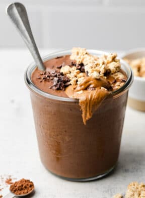 Chocolate protein smoothie in a glass jar topped with granola and nut butter. There is a spoon in the jar as well.