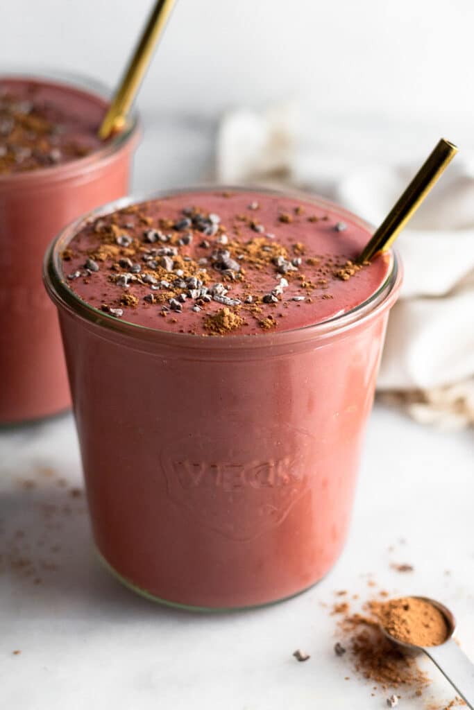 Red smoothie in a glass jar topped with cacao nibs with a straw in it. Behind it is another one.