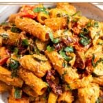 Sun dried tomato chicken pasta in a bowl topped with basil and parmesan cheese. Behind it is another bowl of pasta and bowl of sun-dried tomatoes.