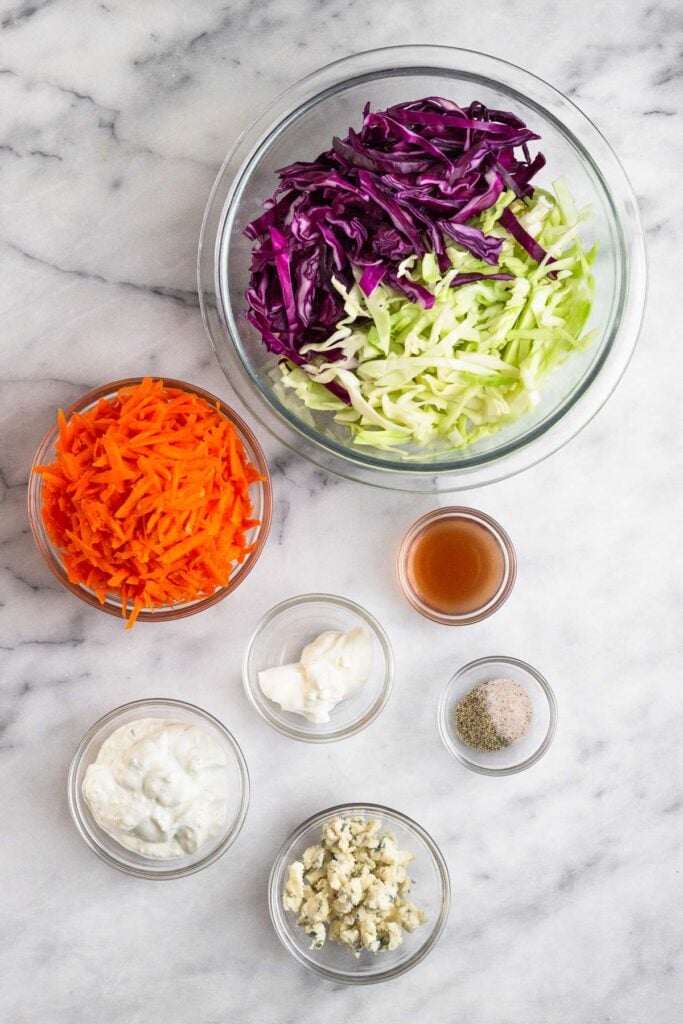 White counter with a bowl of shredded cabbage, bowl of red wine vinegar, bowl of salt and pepper, bowl of blue cheese crumbles, bowl of blue cheese dressing, bowl of sour cream, and a bowl of shredded carrots.