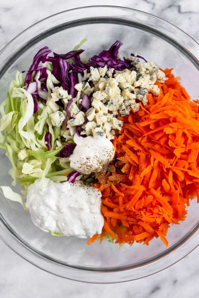 Large glass bowl filled with shredded cabbage, shredded carrots, blue cheese crumbles, sour cream, and blue cheese dressing.