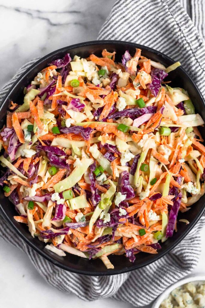 Overhead shot of blue cheese coleslaw in a black bowl. The bowl is on top of a kitchen towel and next to it a bowl of blue cheese.