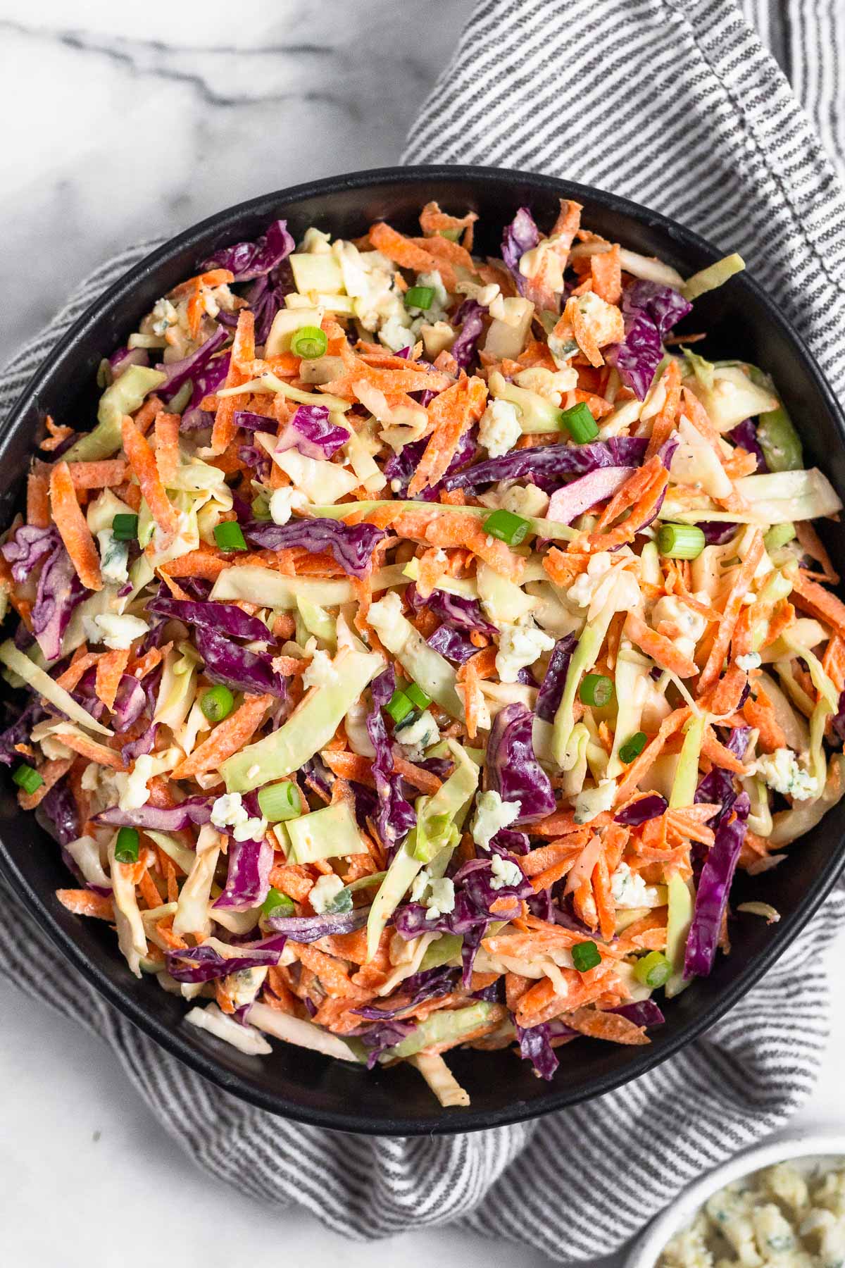 Overhead shot of blue cheese coleslaw in a black bowl. The bowl is on top of a kitchen towel and next to it a bowl of blue cheese.
