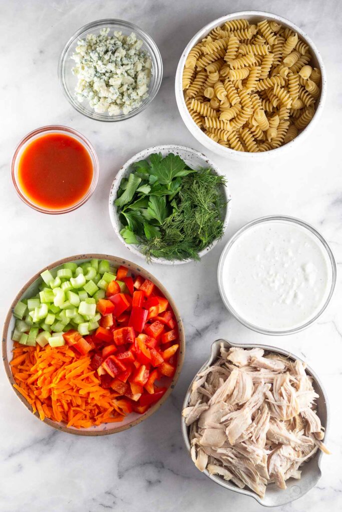 White counter with a bowl of blue cheese crumbles, bowl of dry pasta, plate of fresh herbs, bowl of blue cheese dressing, bowl of shredded chicken, bowl of carrots, peppers, and celery, and a bowl of hot sauce.