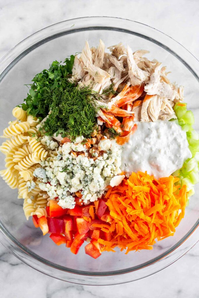 Bowl of cooked pasta, fresh herbs, shredded chicken, hot sauce, blue cheese dressing, celery, carrots, red peppers, and blue cheese crumbles.