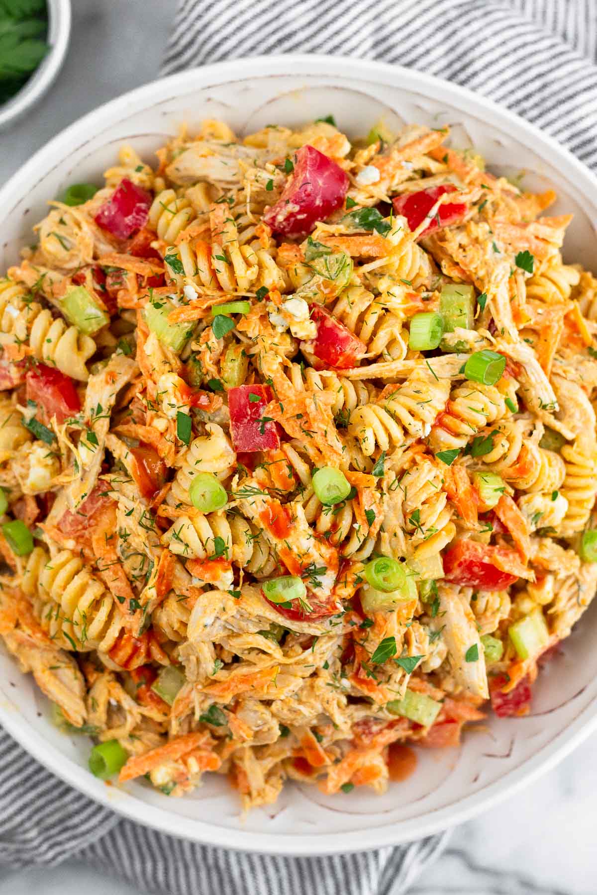 Buffalo chicken pasta salad in a large white bowl. It is on a striped towel and next to it a bowl of herbs.
