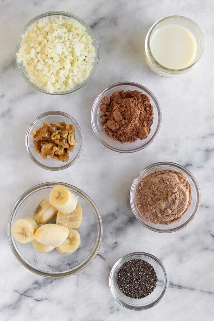 White marble counter with a jar of milk, a bowl of cacao powder, a bowl of chocolate protein powder, a bowl of chia seeds, a bowl of frozen bananas, a bowl of walnuts, and a bowl of frozen cauliflower rice.
