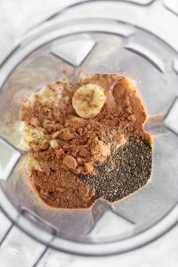 Overhead shot of a blender filled with milk, frozen bananas, chocolate protein powder, cacao powder, and chia seeds.