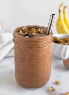 Chunky monkey smoothie in a glass jar with a straw in it. It is topped with chocolate chips and walnuts. Behind the jar are some bananas, a bowl of walnuts, and a spoon of cacao powder.
