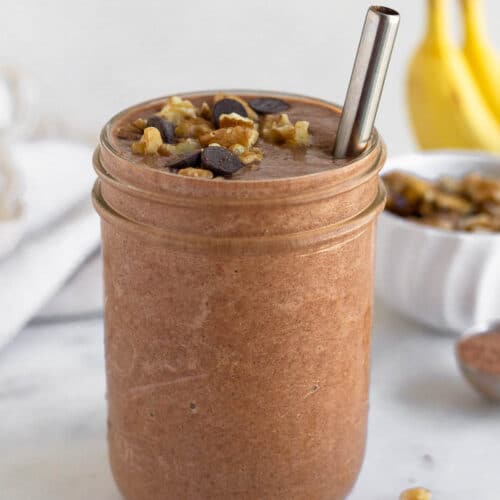 Chunky monkey smoothie in a glass jar with a straw in it. It is topped with chocolate chips and walnuts. Behind the jar are some bananas, a bowl of walnuts, and a spoon of cacao powder.