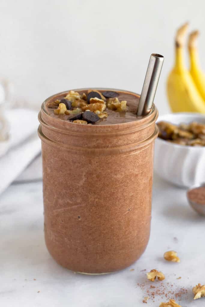 Chunky monkey smoothie in a glass jar with a straw in it. It is topped with chocolate chips and walnuts. Behind the jar are some bananas, a bowl of walnuts, and a spoon of cacao powder.