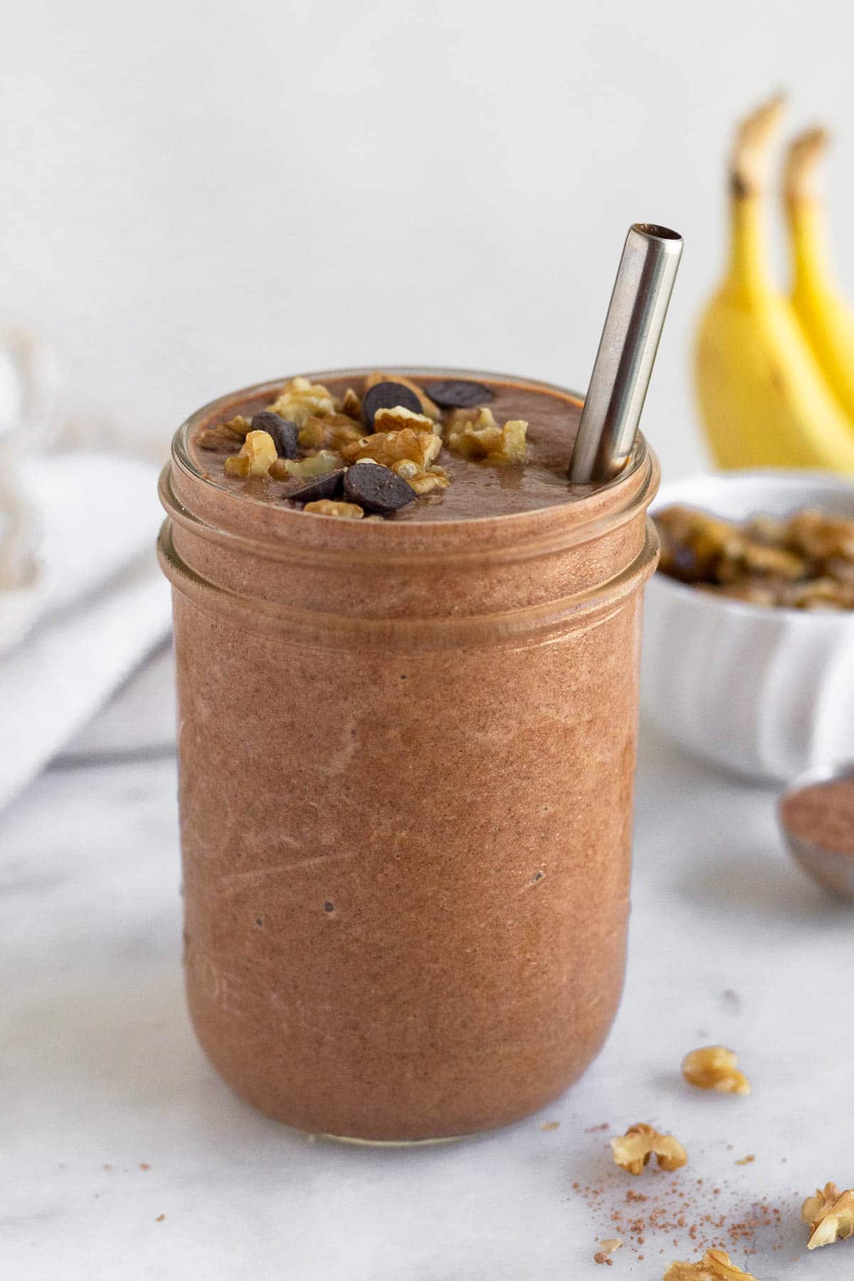 Chunky monkey smoothie in a glass jar with a straw in it. It is topped with chocolate chips and walnuts. Behind the jar are some bananas, a bowl of walnuts, and a spoon of cacao powder.