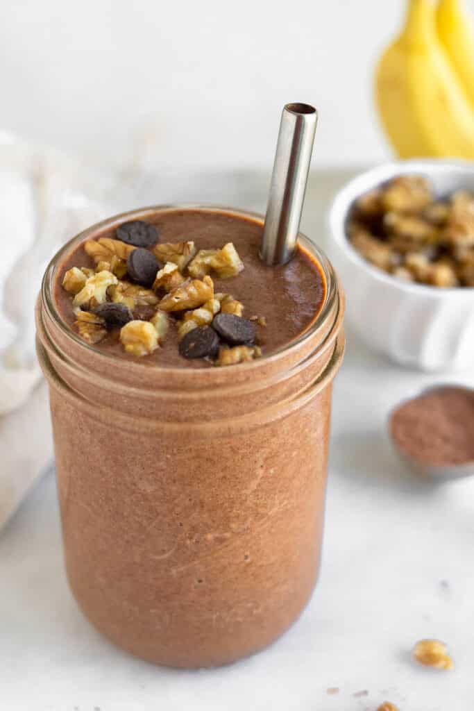 Chunky monkey protein smoothie in a jar topped with walnuts and chocolate chips with a metal straw in it. Behind it is a spoon full of cacao powder, a dish of walnuts, and some bananas.