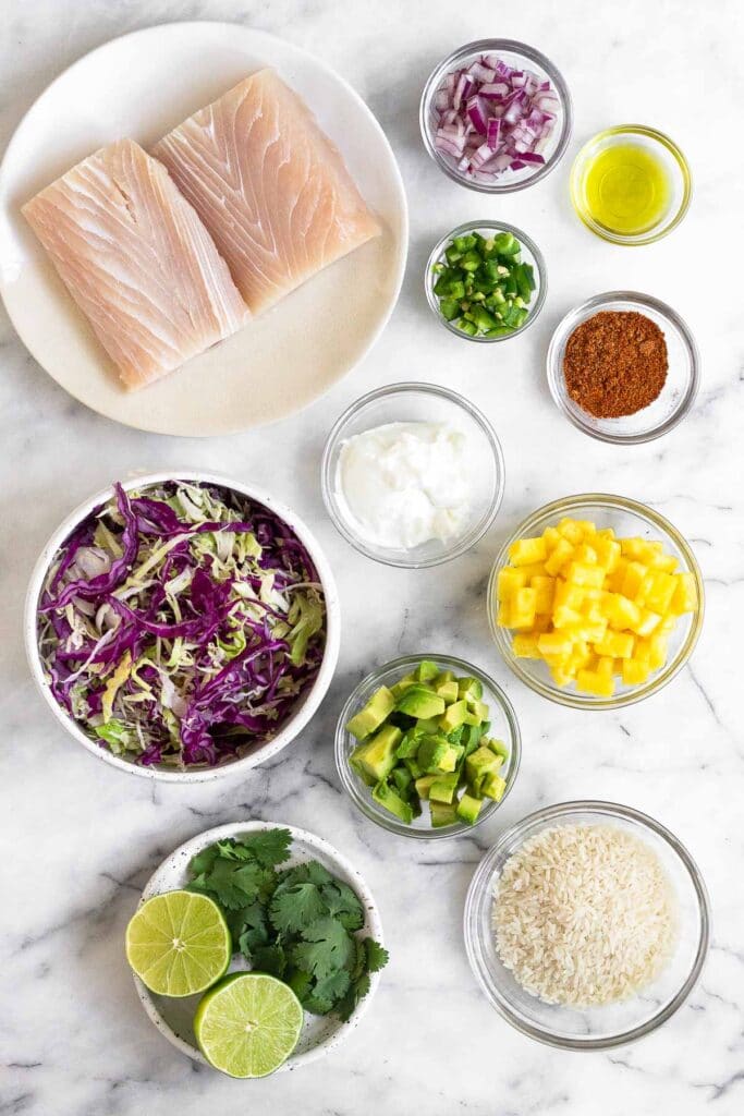 Overhead shot a plate with 2 filets of white fish, bowl of red onion, bowl of oil, bowl of taco seasoning, bowl of diced jalapeño, bowl of diced pineapple, bowl of sour cream, bowl of uncooked white rice, bowl of dice avocado, plate of limes and cilantro, and a bowl of shredded cabbage.