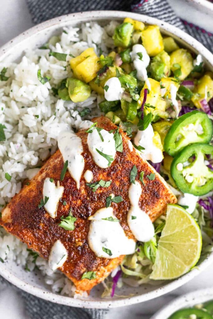 Overhead shot of a fish taco bowl with white rice, pineapple salsa, cabbage, blackened fish, and drizzled with white sauce. It is garnished with jalapeños and a lime wedge.