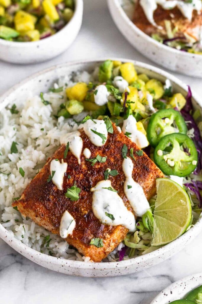 Fish taco bowls with rice, pineapple salsa, cabbage, and blackened fish drizzled with a white sauce. Behind it is another bowl and a bowl of pineapple salsa.