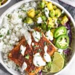 Fish taco bowl with cilantro white rice, pineapple salsa, cabbage, seasoned fish, and white sauce in a bowl. Around it is a plate of jalapeños, a bowl of salsa, and another fish taco bowl.