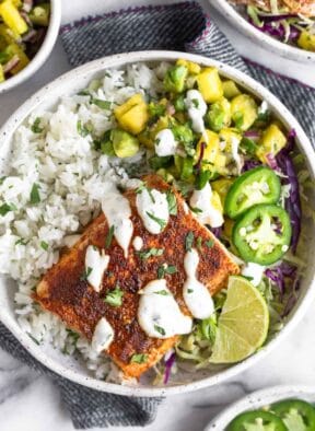 Fish taco bowl with cilantro white rice, pineapple salsa, cabbage, seasoned fish, and white sauce in a bowl. Around it is a plate of jalapeños, a bowl of salsa, and another fish taco bowl.