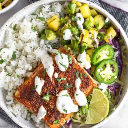 Fish taco bowl with cilantro white rice, pineapple salsa, cabbage, seasoned fish, and white sauce in a bowl. Around it is a plate of jalapeños, a bowl of salsa, and another fish taco bowl.