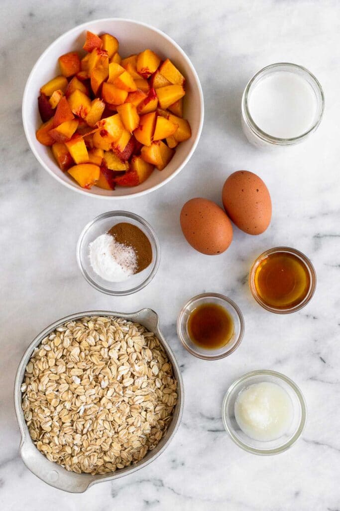 White counter top with a bowl of diced peaches, jar of milk, 2 eggs, bowl of maple syrup, bowl of coconut oil, bowl of rolled oats, bowl of vanilla extract, and bowl of spices.