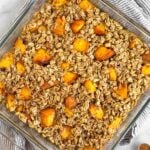 Peach baked oatmeal in a glass dish. It is sitting on a kitchen towel with half a peach next to it.