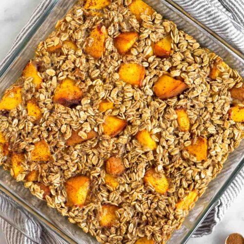Peach baked oatmeal in a glass dish. It is sitting on a kitchen towel with half a peach next to it.