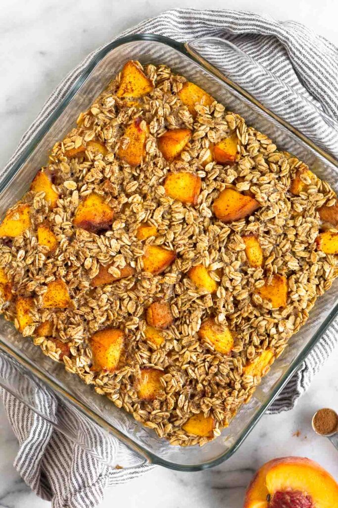 Peach baked oatmeal in a glass dish. It is sitting on a kitchen towel with half a peach next to it.