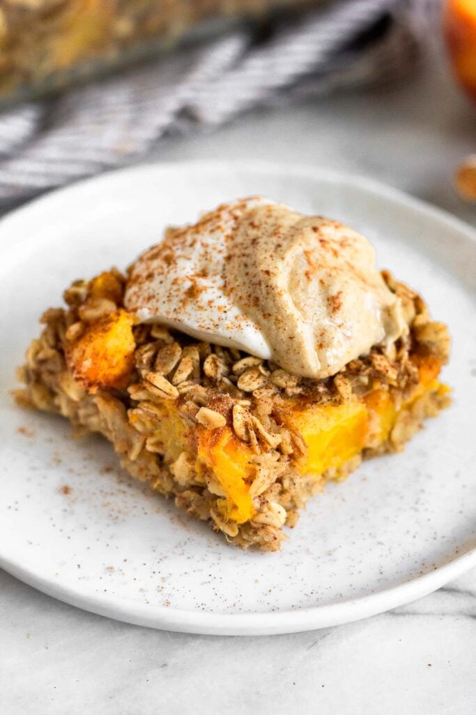 A slice of peach baked oatmeal on a small plate. It is topped with yogurt, nut butter, and cinnamon.