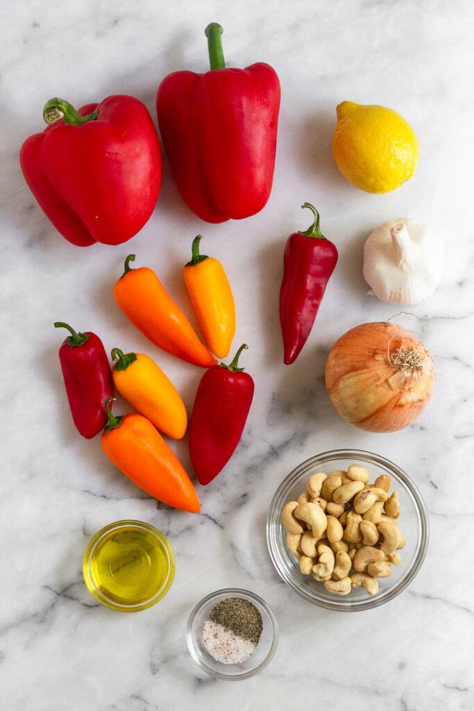 Overhead shot with red pepper peppers, a lemon, bulb of garlic, an onion, bowl of cashews, bowl of salt and pepper, bowl of olive oil, mini bell peppers, and a Fresno pepper.