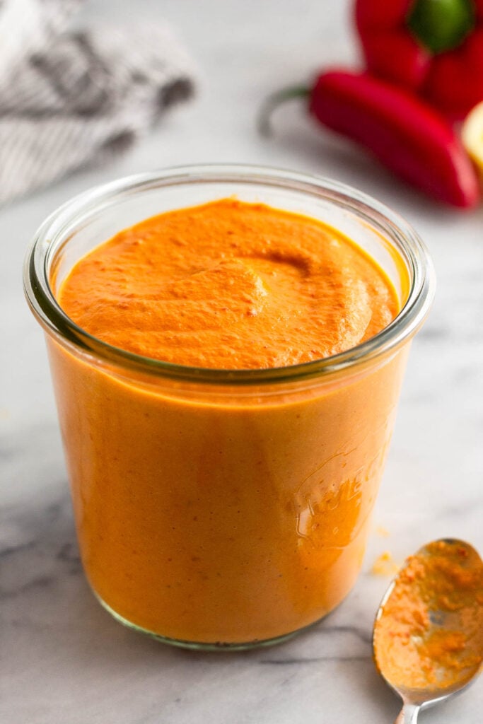 Spicy red pepper sauce in a jar. Next to it a spoon with more sauce on it and behind the jar are peppers and a kitchen towel.