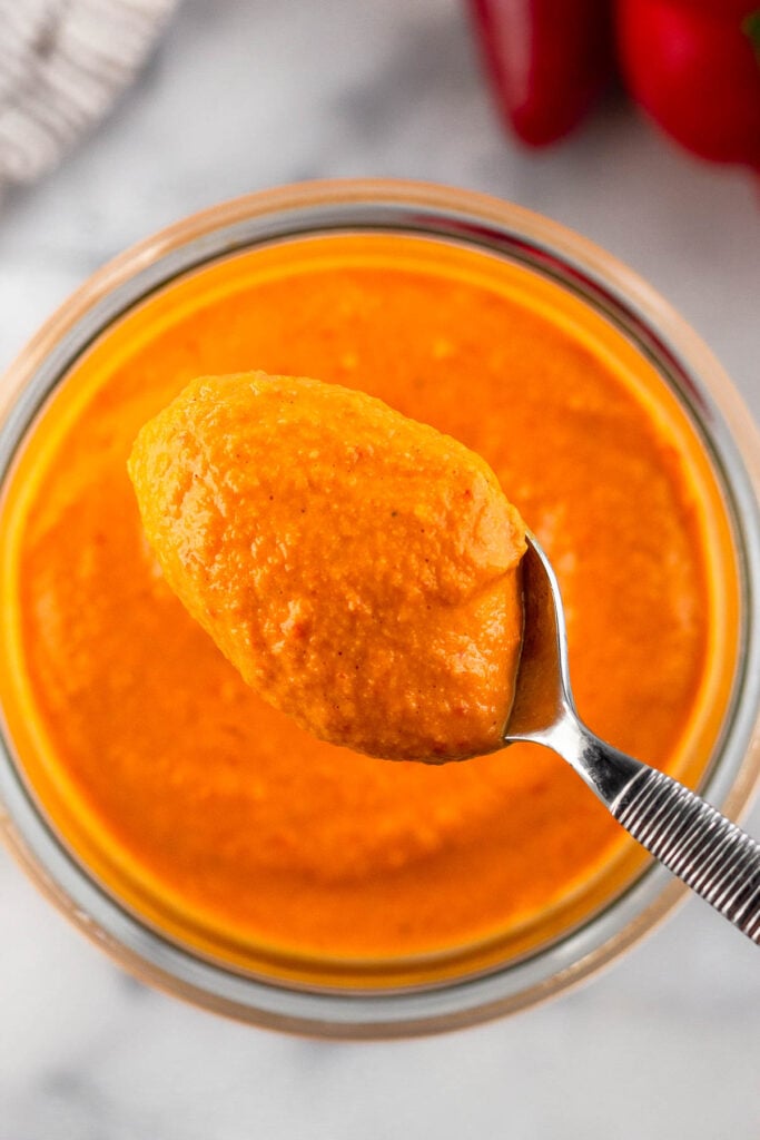 Overhead shot of creamy red pepper sauce with a spoon filled with it overtop of the jar.