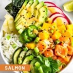 Salmon poke bowl with salmon, rice, veggies, mango, and avocado drizzled with spicy mayo. Behind it is another bowl, more sesame seeds, lime wedges, and a bowl of soy sauce is in front of it.