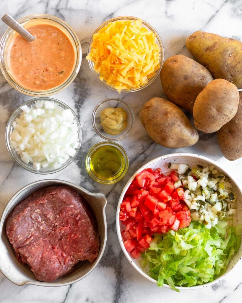 Overhead shot of a jar of special sauce, bowl of shredded cheese, 5 russet potatoes, bowl of tomatoes, lettuce, and pickles, bowl of raw ground beef, bowl of oil, bowl of mustard, and a bowl of diced onion.
