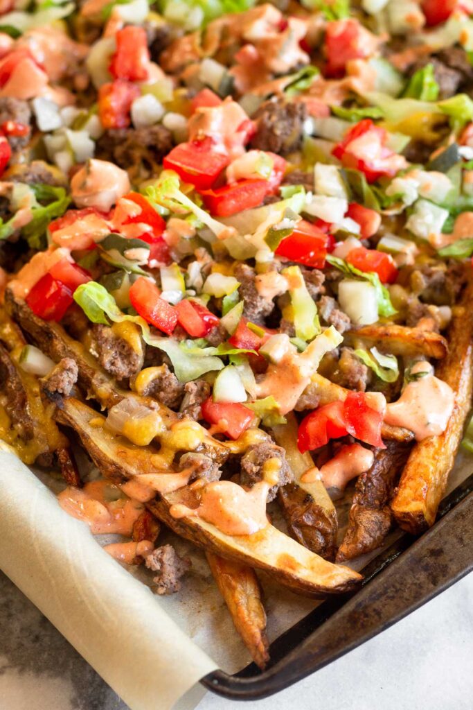 Close up of cheeseburger french fries on a large baking sheet. The fries are topped with ground meat, melted cheese, lettuce, tomato, pickles, and special sauce.