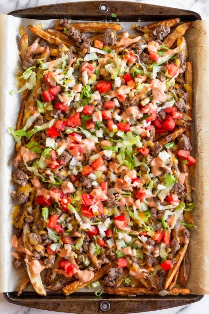 Overhead shot of burger fries on a large baking sheet with melted cheese, ground beef, lettuce, tomato, pickles, and special sauce.