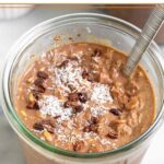 Jar of chocolate coconut overnight oats topped with cacao nibs and shredded coconut. There is spoon in the jar and behind it is another jar and a bowl of coconut.