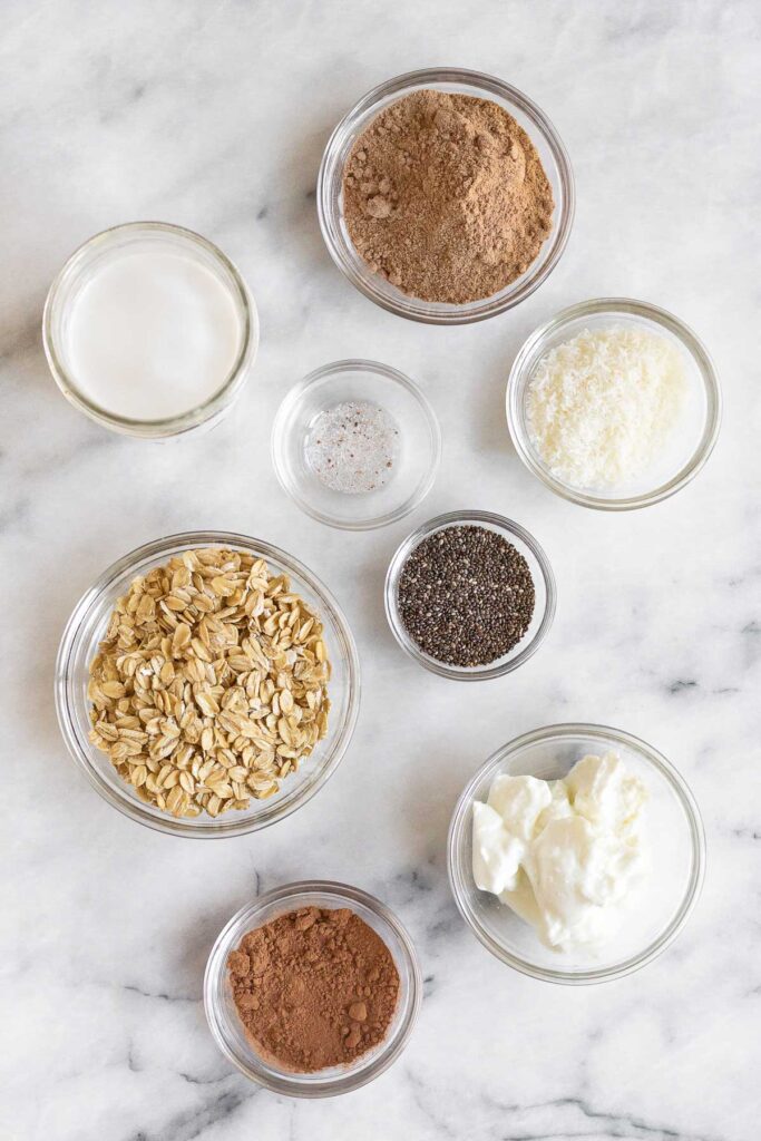 Overhead shot of a white counter with a bowl of chocolate protein powder, bowl of shredded coconut, bowl of chia seeds, bowl of greek yogurt, bowl of cacao powder, bowl of rolled oats, bowl of salt, and a jar of coconut milk.
