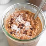 Jar of chocolate coconut overnight oats topped with cacao nibs and shredded coconut. There is spoon in the jar and behind it is another jar and a bowl of coconut.