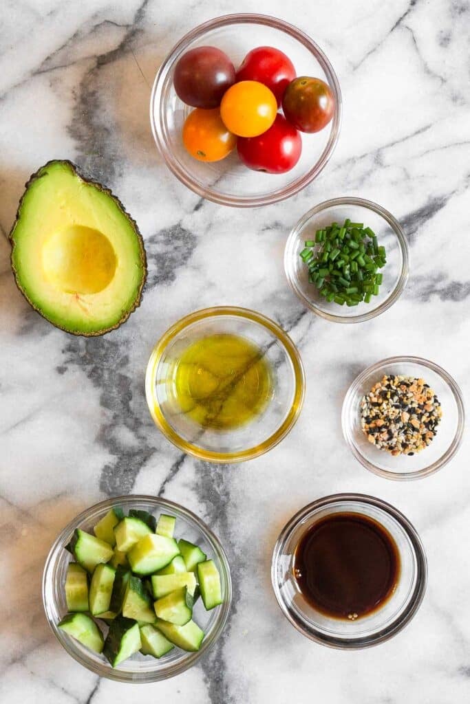 White marble counter with a bowl of cherry tomatoes, a bowl of chopped chives, a bowl of bagel seasoning, a bowl of balsamic vinegar, a bowl of diced cucumbers, a bowl of olive oil, and half an avocado.