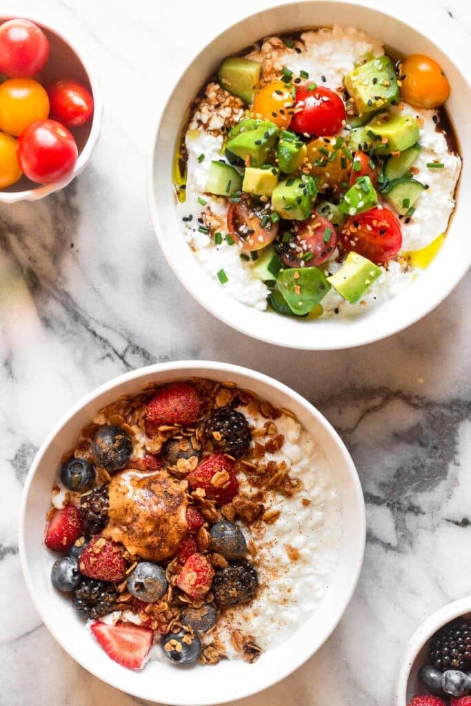 Two white bowls filled with cottage cheese. One is topped with fruit, granola, and peanut butter and the other is topped with tomatoes, cucumbers, avocado, and bagel seasoning. Off to the side around them is a bowl of tomatoes and a bowl of berries.