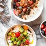 Overhead shot of two cottage cheese bowls. One is sweet with berries, granola, and peanut butter and the other is savory with tomato, cucumber, avocado, and bagel seasoning. Next to the bowls is a kitchen towel, a bowl of berries, and a bowl of cherry tomatoes.
