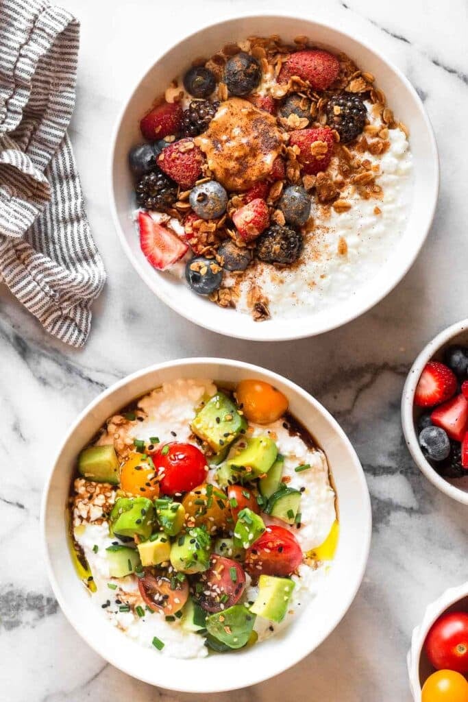 Overhead shot of two cottage cheese bowls. One is sweet with berries, granola, and peanut butter and the other is savory with tomato, cucumber, avocado, and bagel seasoning. Next to the bowls is a kitchen towel, a bowl of berries, and a bowl of cherry tomatoes.