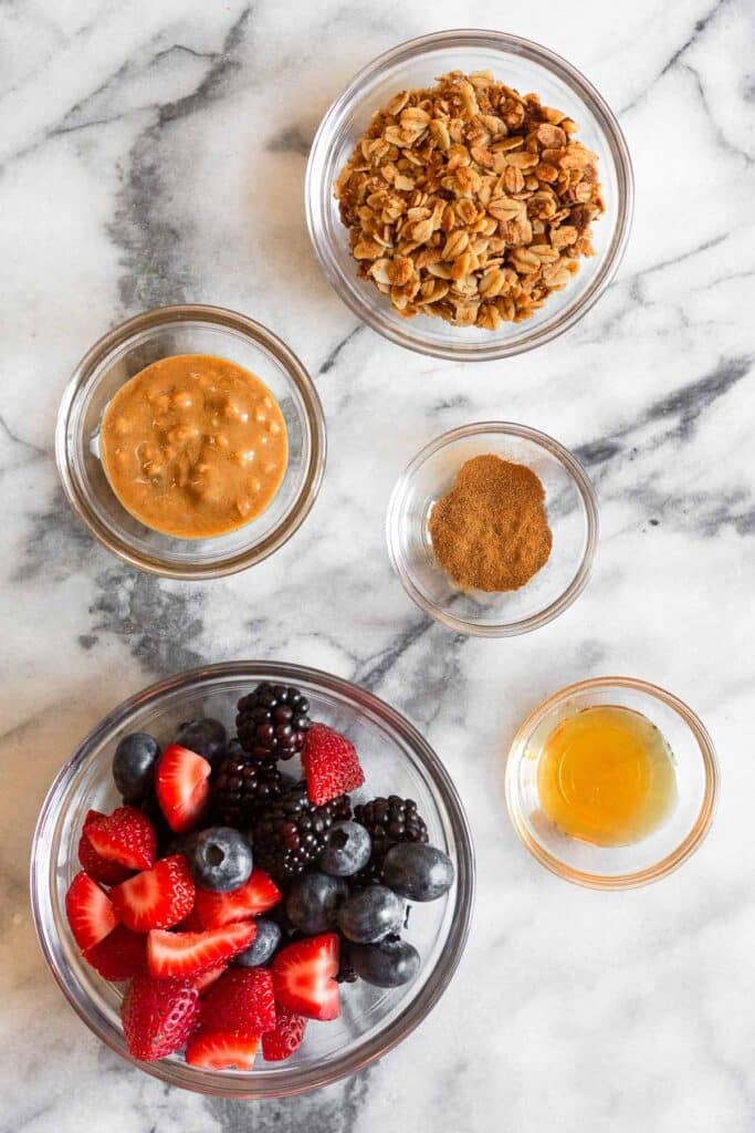 White marble counter with a bowl of granola, a bowl of cinnamon, a bowl of honey, a bowl of mixed berries, and bowl of peanut butter.