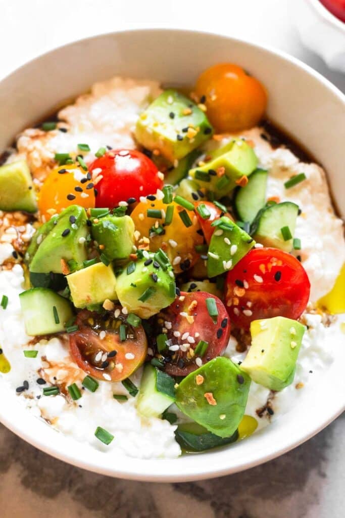 A close-up of savory cottage cheese bowl topped with sliced cherry tomatoes, diced cucumber, and diced avocado. It is topped with olive oil, balsamic vinegar, chopped chives, and everything bagel seasoning.