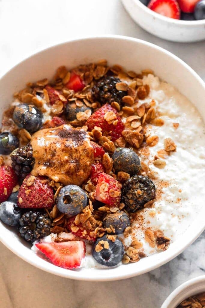 A close-up of a cottage cheese breakfast bowl topped with strawberries, blueberries, blackberries, granola, peanut butter, honey, and sprinkled with cinnamon. In front of the bowl is a small dish of granola and behind the bowl is a bowl of berries.