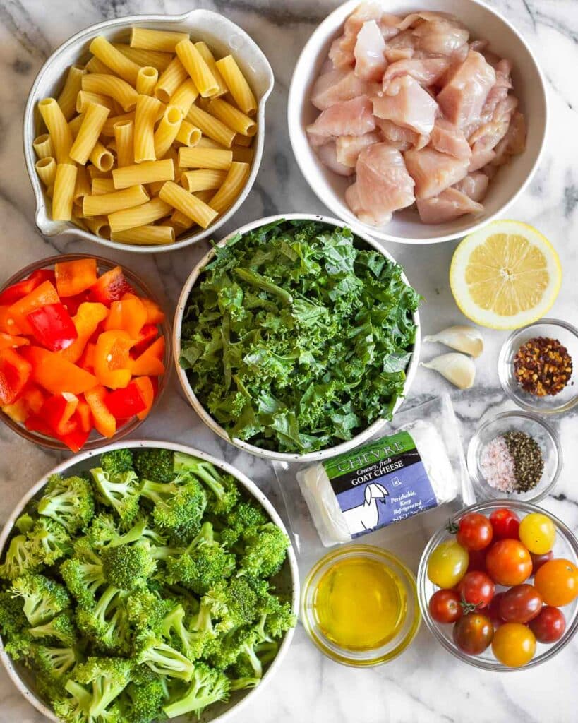 Overhead shot of a bowl of dry pasta, bowl of diced raw chicken, half a lemon, bowl of red pepper flakes, 2 garlic cloves, bowl of salt and pepper, bowl of cherry tomatoes, bowl of olive oil, package of goat cheese, bowl of broccoli florets, bowl of chopped kale, and a bowl of red pepper flakes.