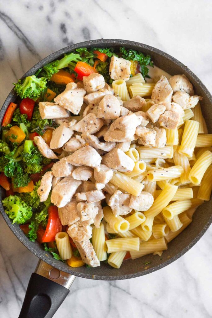 Large saute pan filled with sautéed veggies, cooked chicken, cooked pasta, and some sauce.