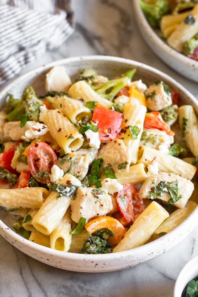 Pasta with goat cheese, chicken, and veggies in a bowl. Behind it is another bowl and a kitchen towel.