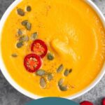 Overhead shot of Instant Pot pumpkin soup garnished with pumpkin seeds, chili pepper, and pepper. Next to it is more sliced peppers and another bowl.